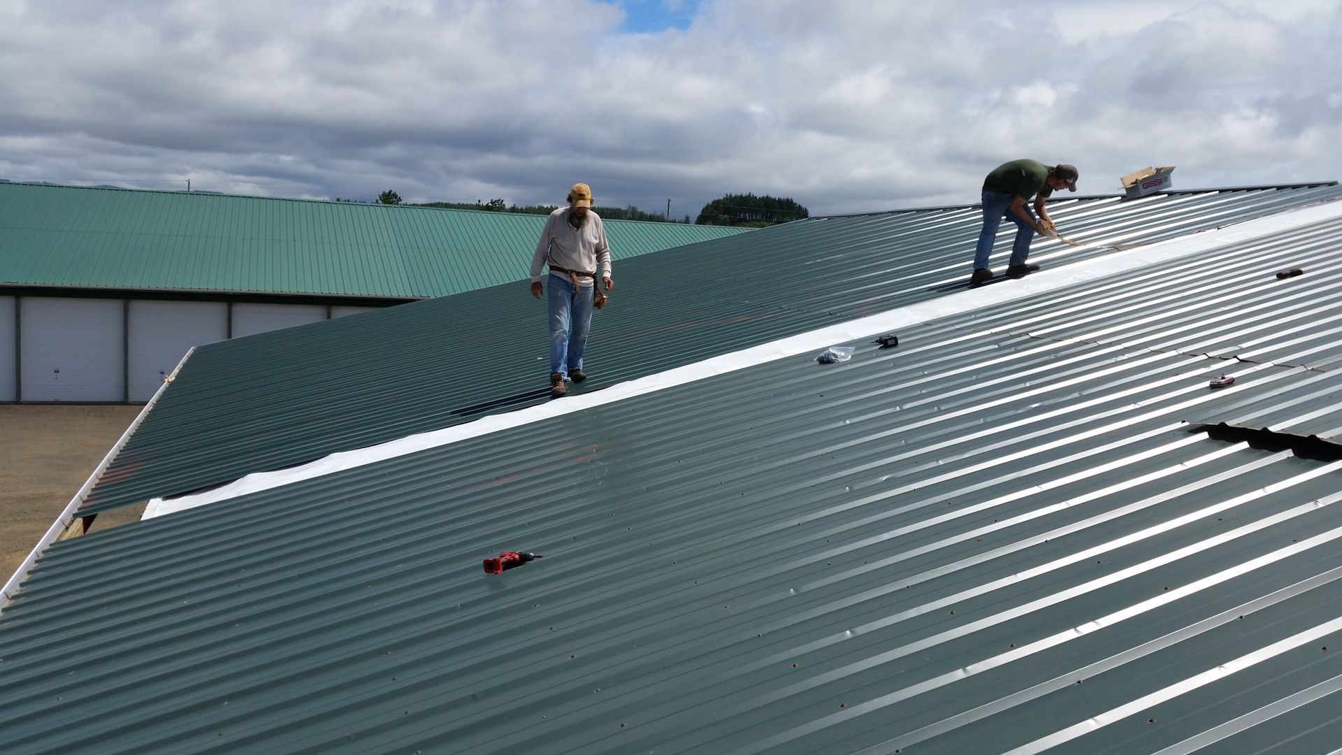 Workers on a green metal roof, one standing and one crouching near the ridge under a cloudy sky.