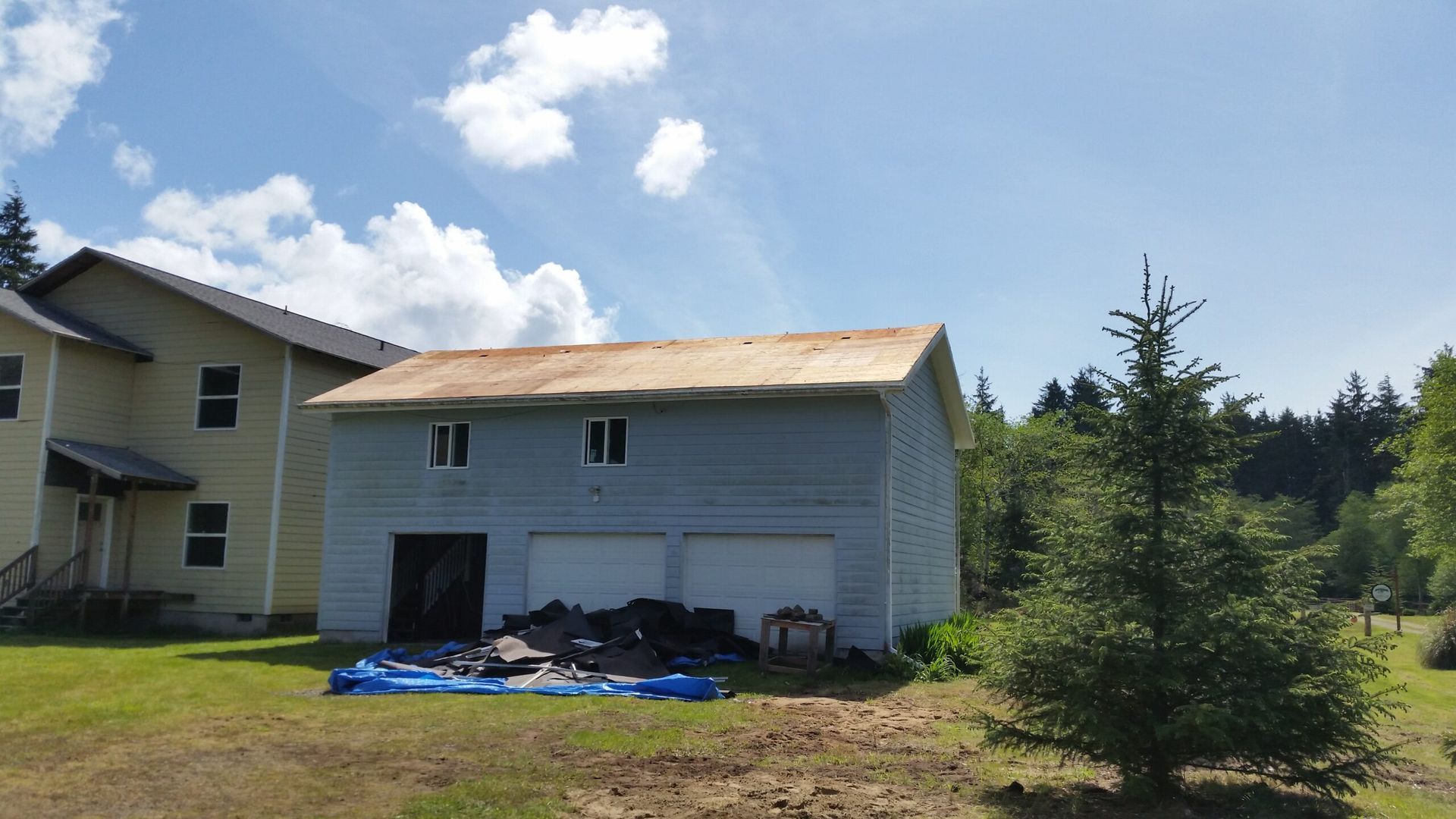 Small white outbuilding with peeling roof, trash by the door, beside a house and tree in a grassy yard.