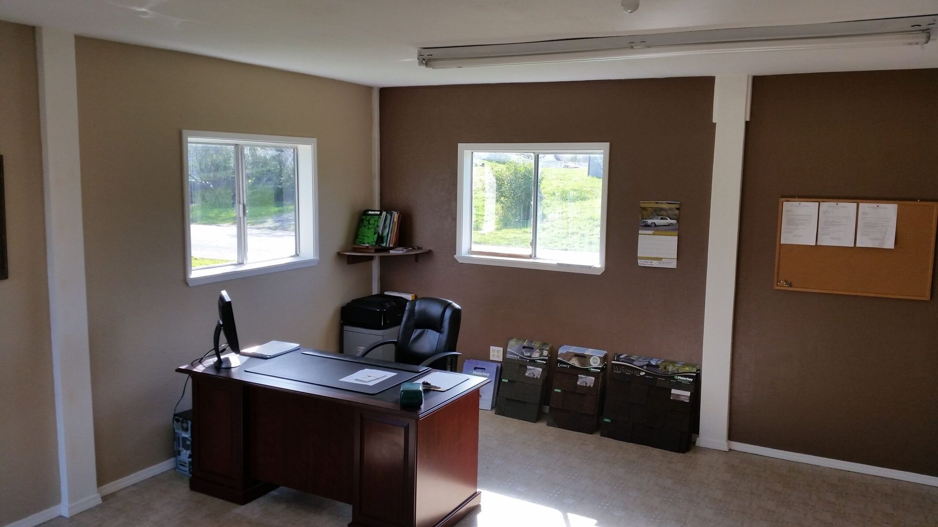 Small office with a desk, chair, windows, brown walls, and filing boxes along the floor.
