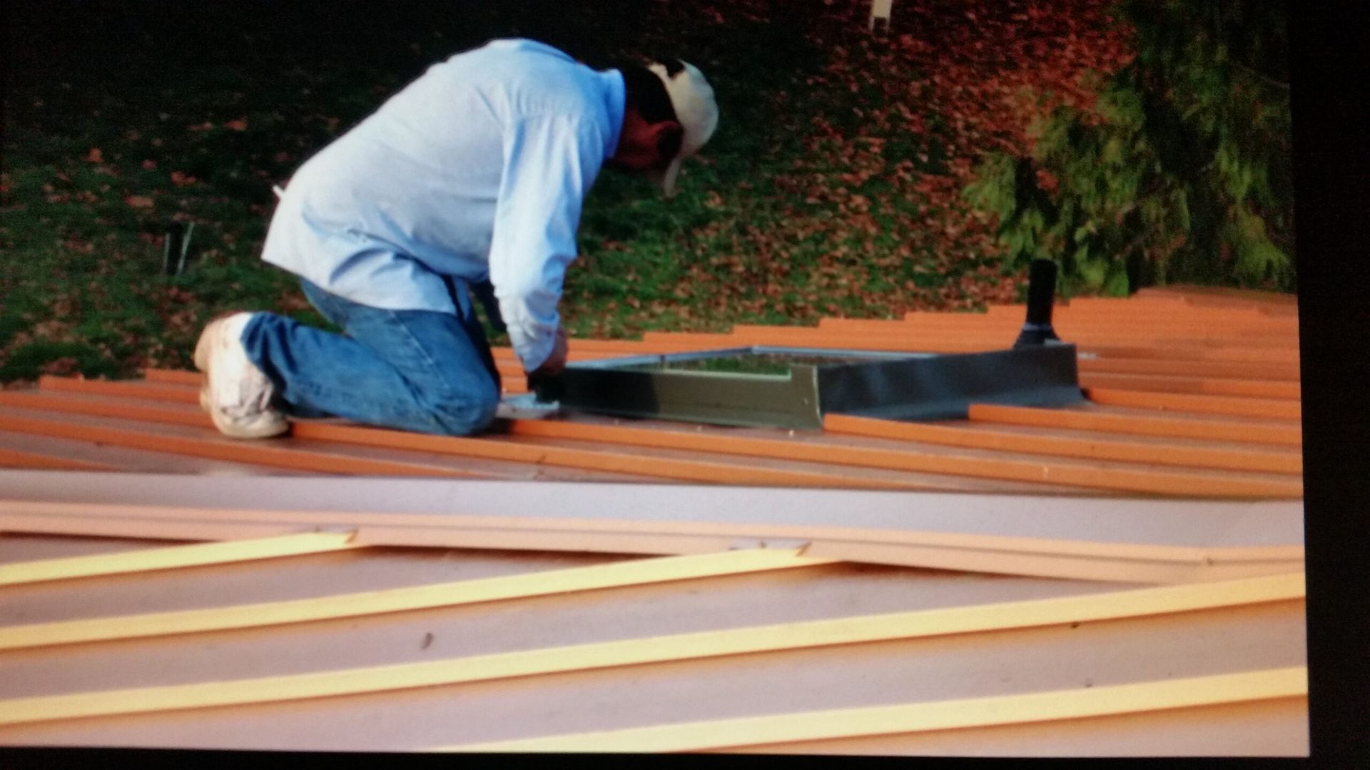 Worker kneeling on a roof, installing black flashing over orange roofing panels.