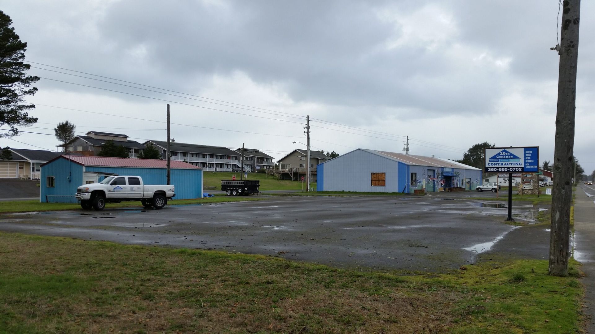Small blue buildings and parked vehicles on a wet rural roadside under cloudy skies.