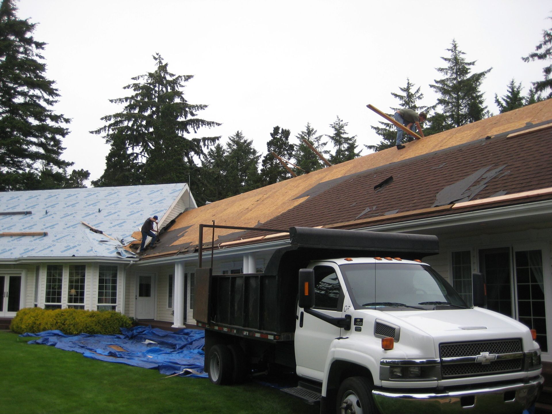 White dump truck beside a house with roof damage and blue tarp during repairs