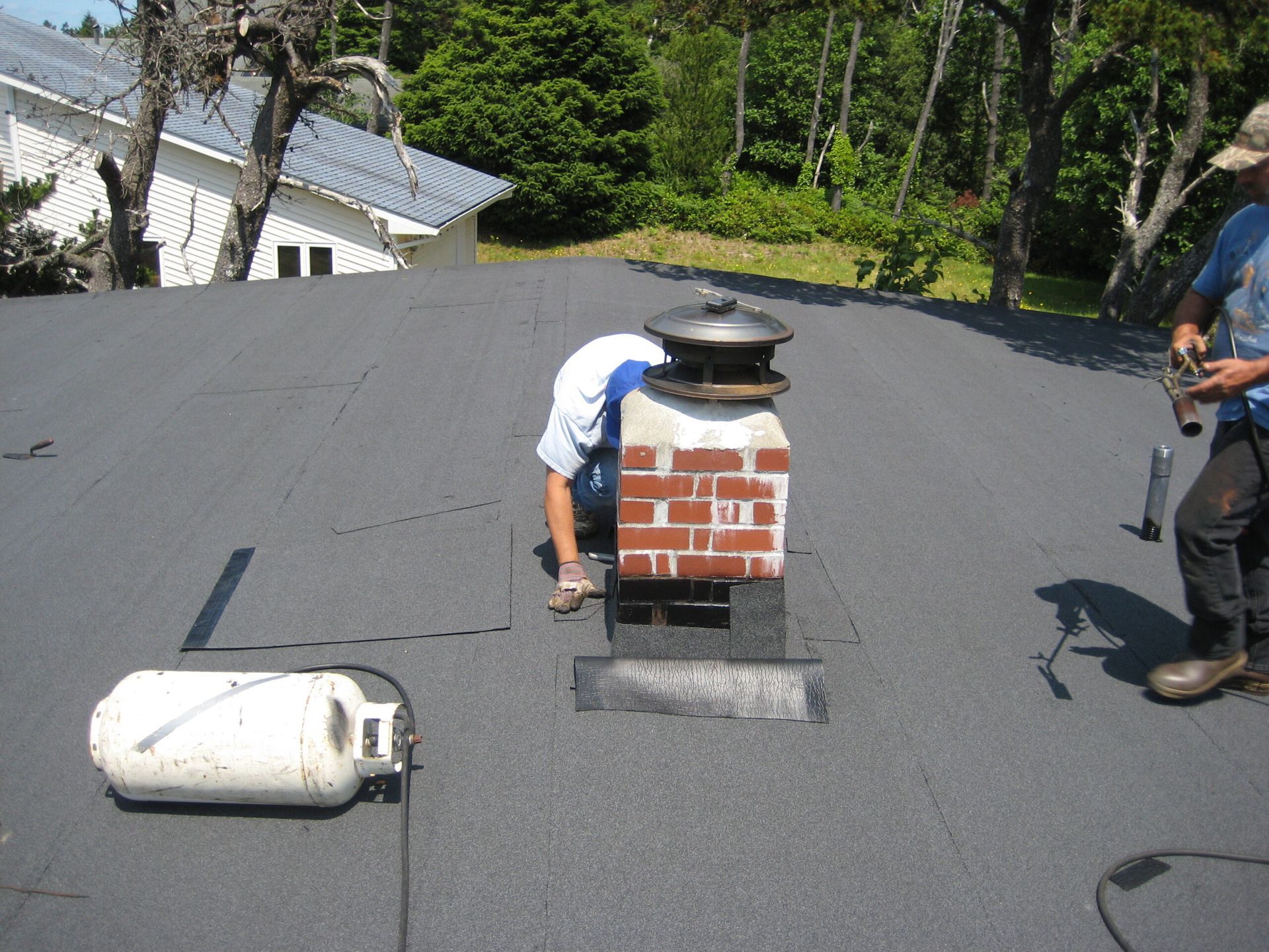 Worker repairing roof vent on flat gray roof, with tools and white insulation roll nearby.