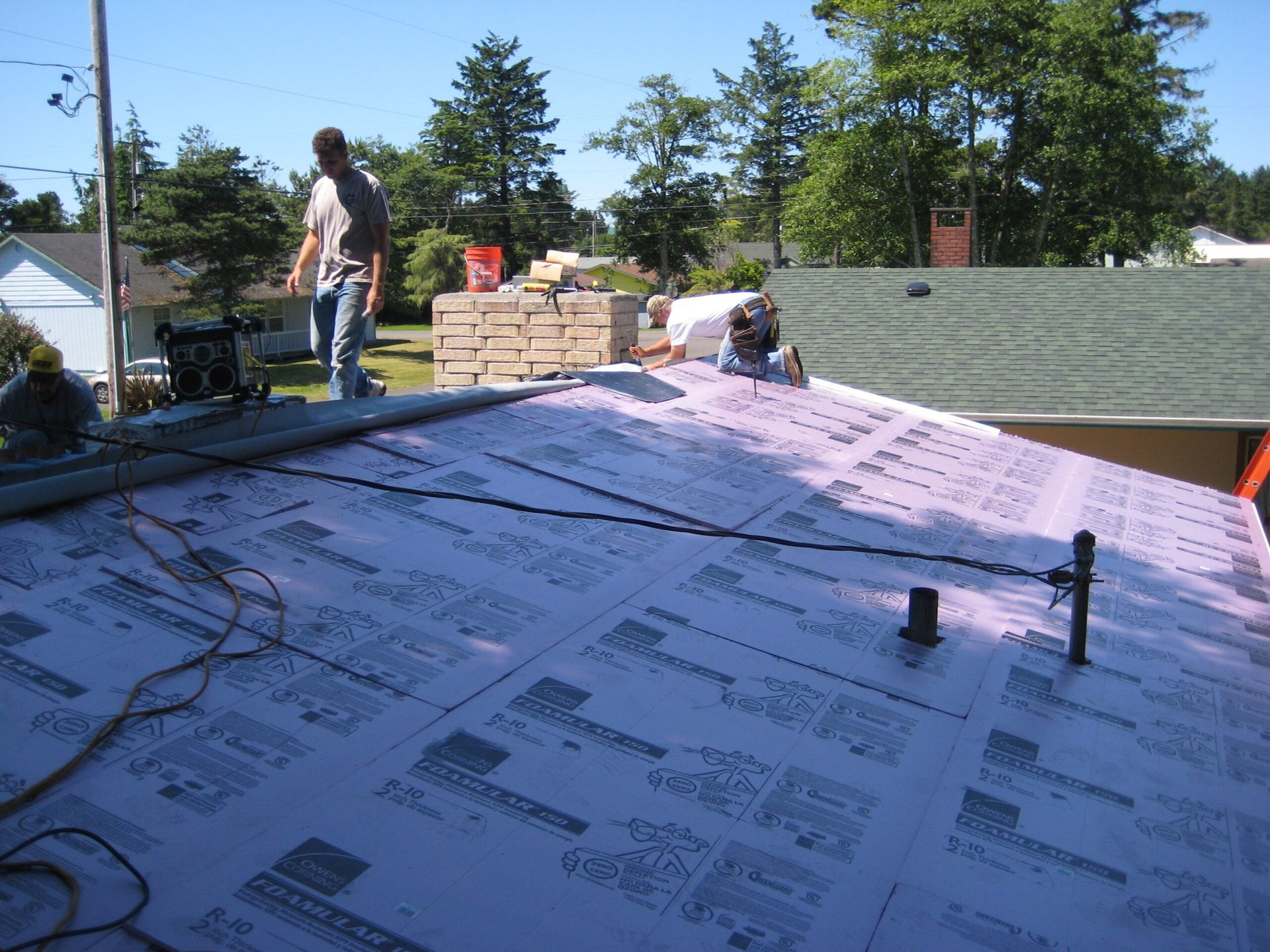Roofers installing shingles on a sloped roof with pink underlayment and stacks of materials nearby.