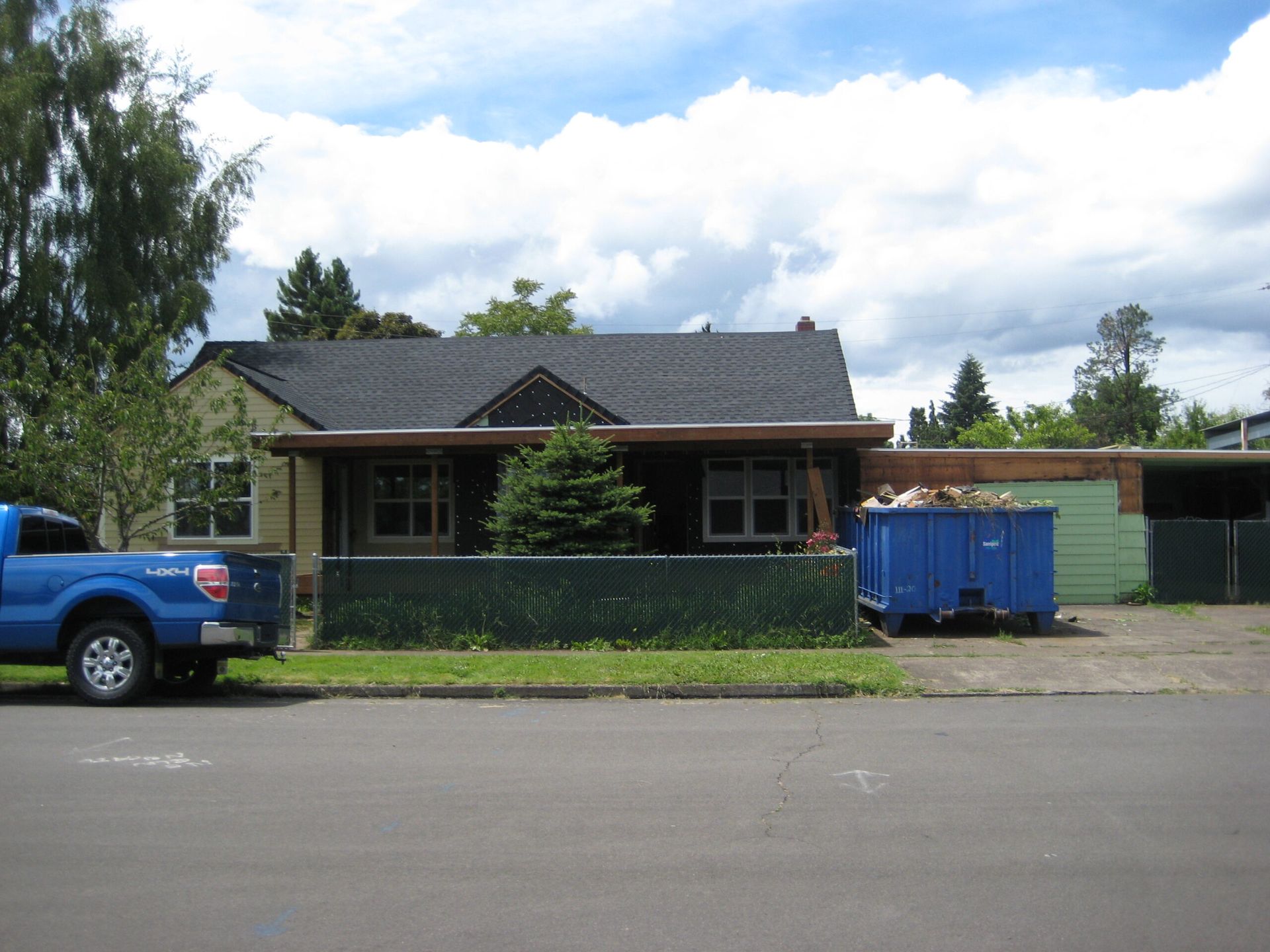 Small house with a dark roof, blue pickup truck, and two blue bins by the roadside under a cloudy sky.