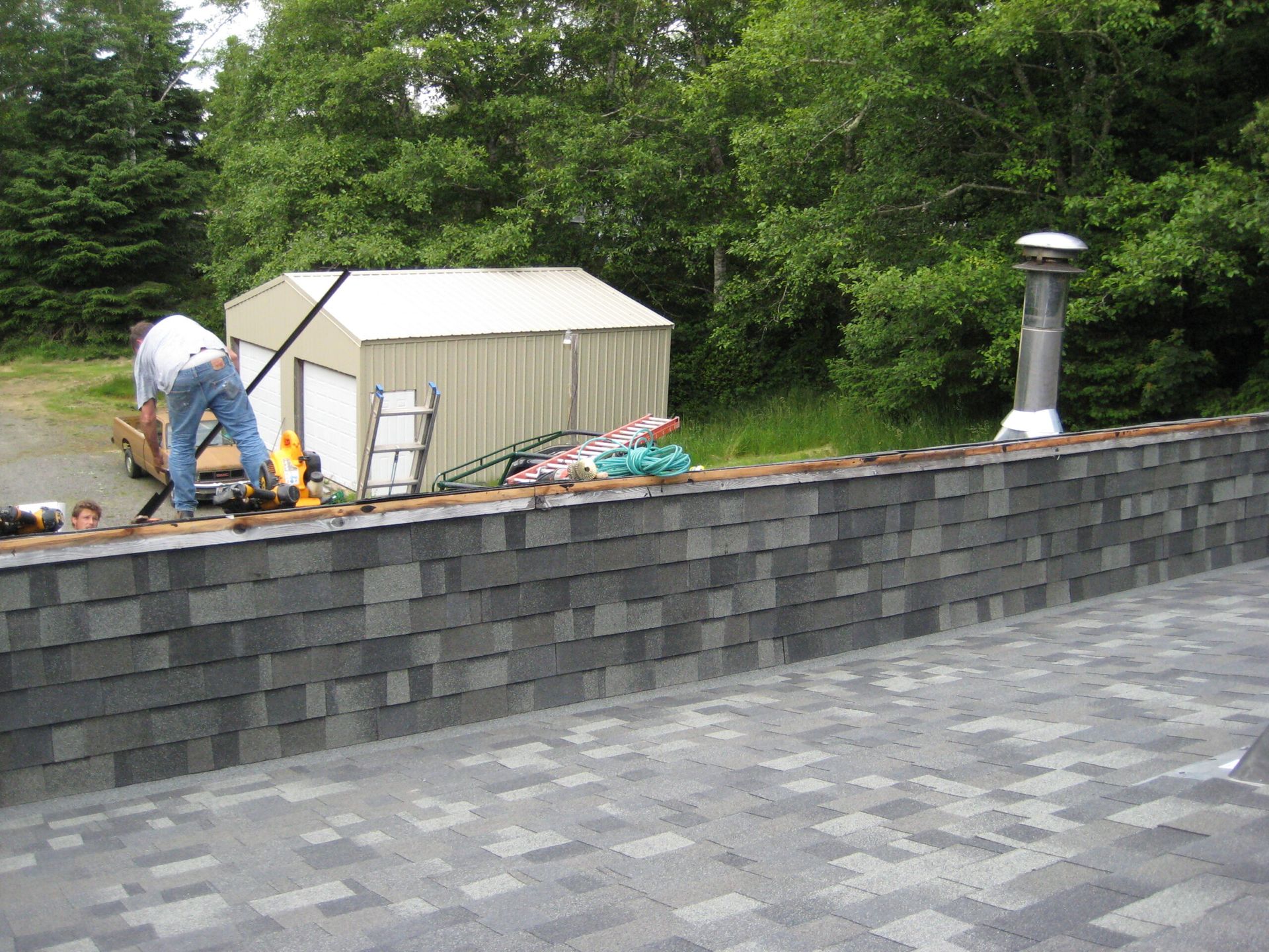 Workers on a rooftop near a white shed, with a gray shingle roof and green trees in the background