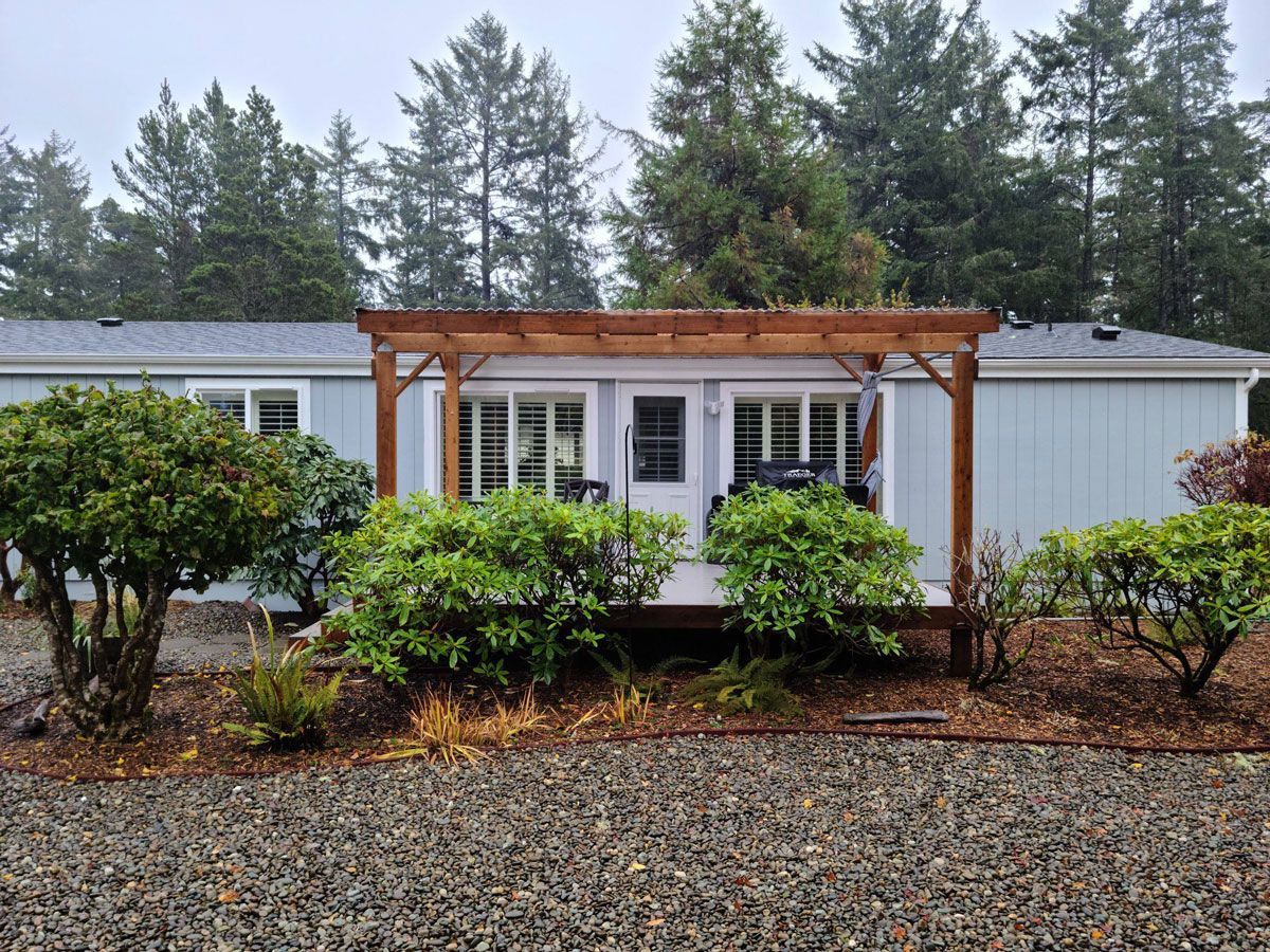 Small white mobile home with a wooden pergola, front shrubs, and trees in a wooded yard.