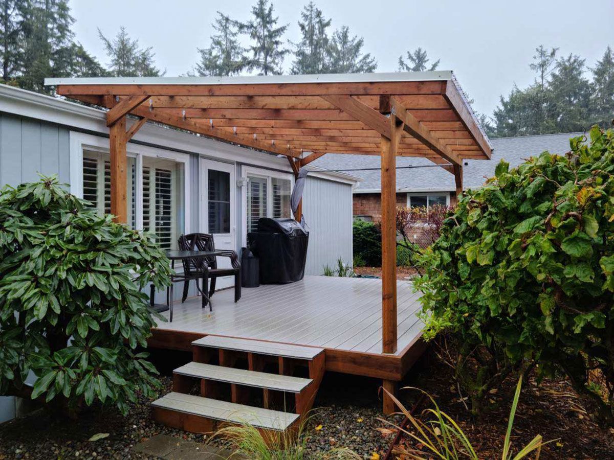 Wooden deck with pergola beside a white house, surrounded by lush green shrubs and trees.