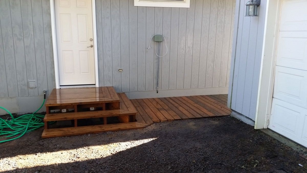 Small wooden steps and a doormat outside a white door beside a gray wall and coiled green hose.