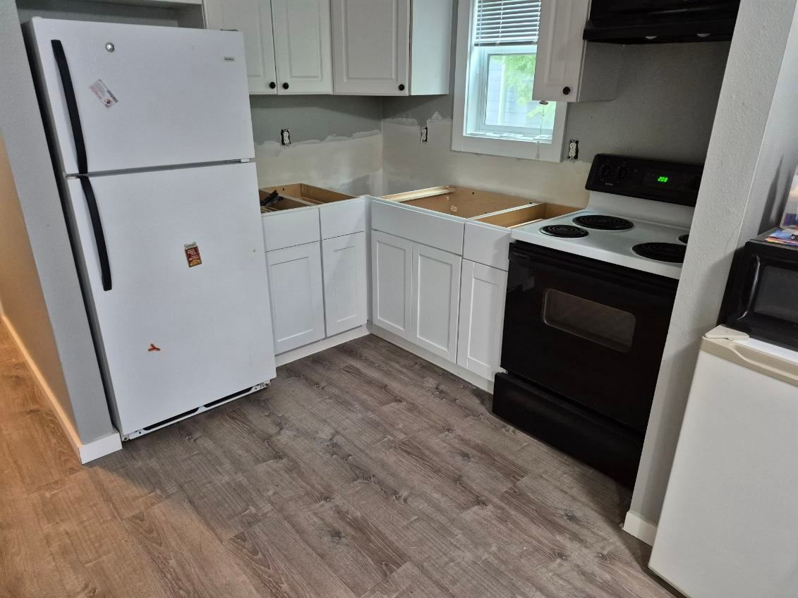 Small white kitchen with fridge, stove, cabinets, and wood-look flooring.