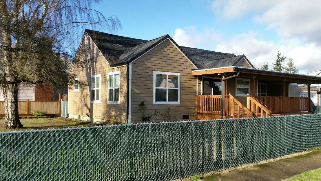 Single-story beige house with porch and black roof behind a green fence under a blue sky