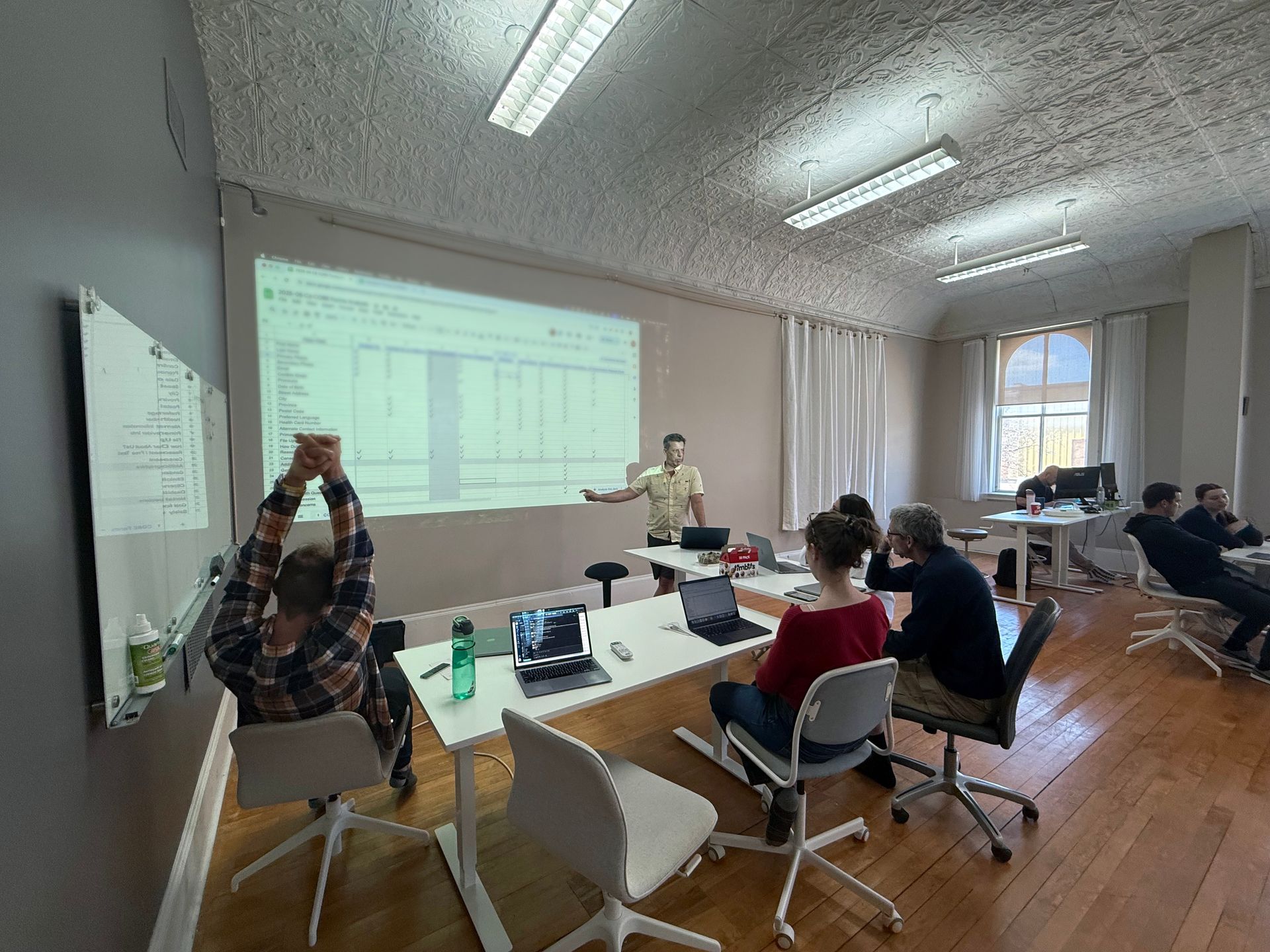 A presentation in a bright room: A man points at a screen, while several people watch.