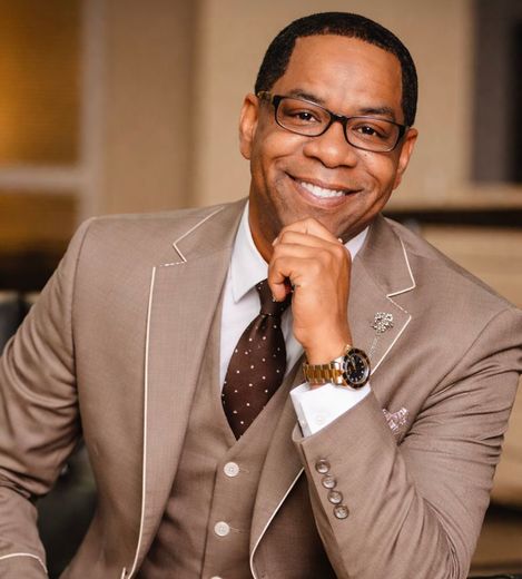 Dr. Marlon Moore in a tan suit and glasses, resting his chin on his hand indoors