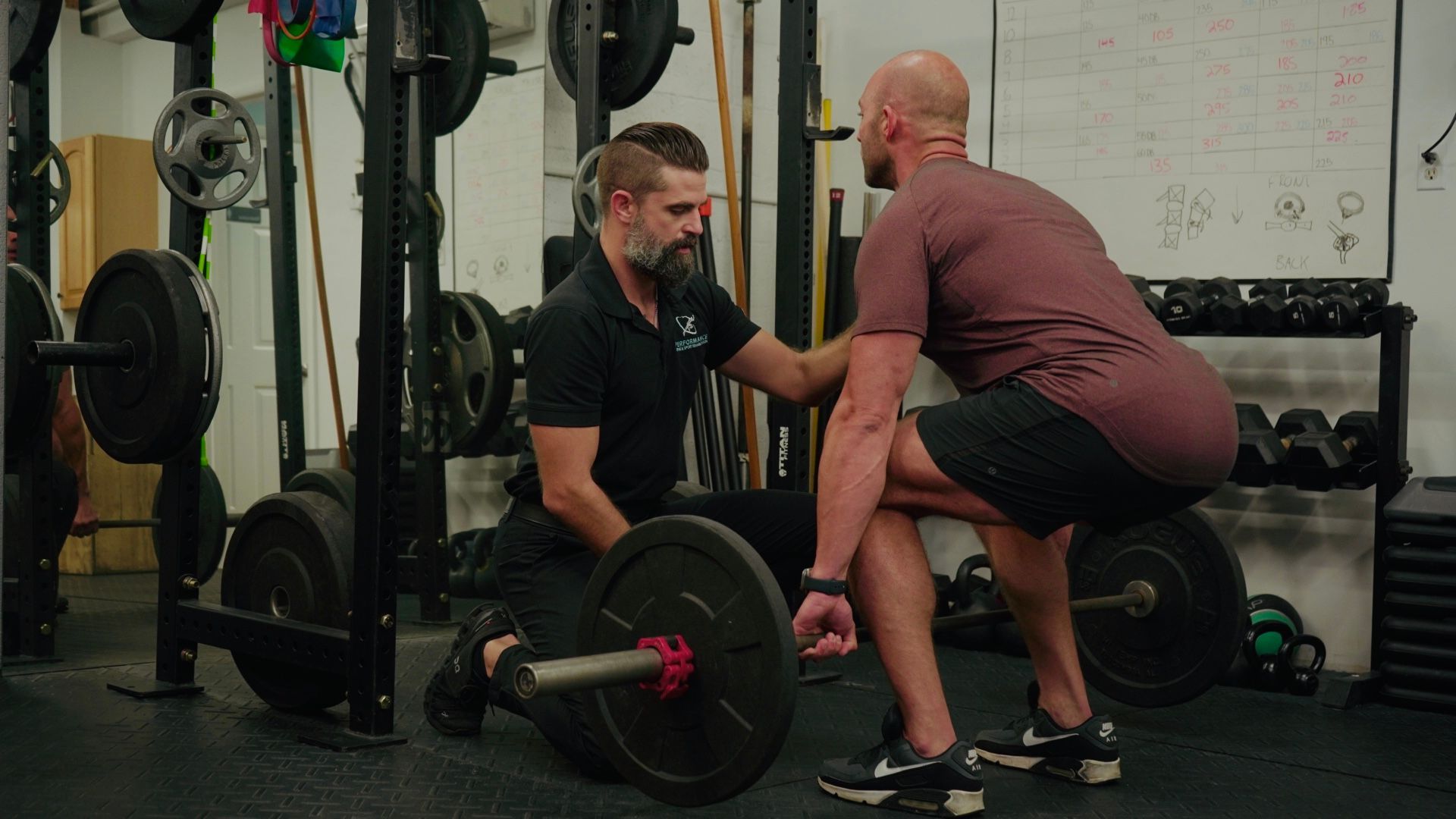 Man assisting another man with a barbell deadlift in a gym.