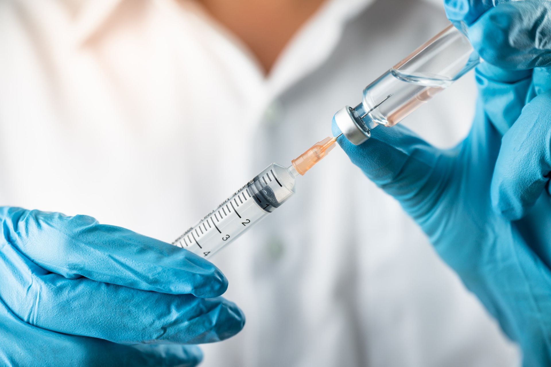 Person in blue gloves drawing liquid from a vial into a syringe.