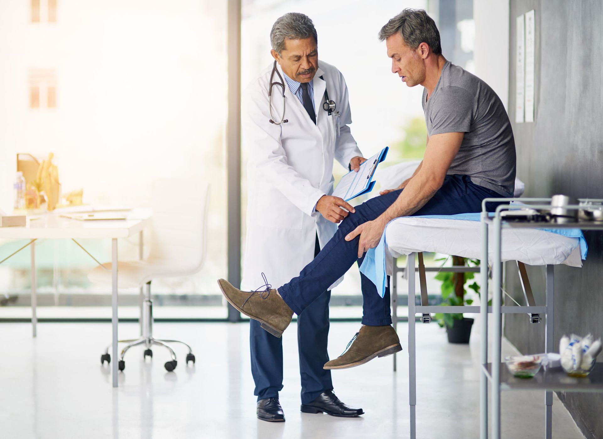 Doctor examining a patient's knee in an office; sunlight streams in.