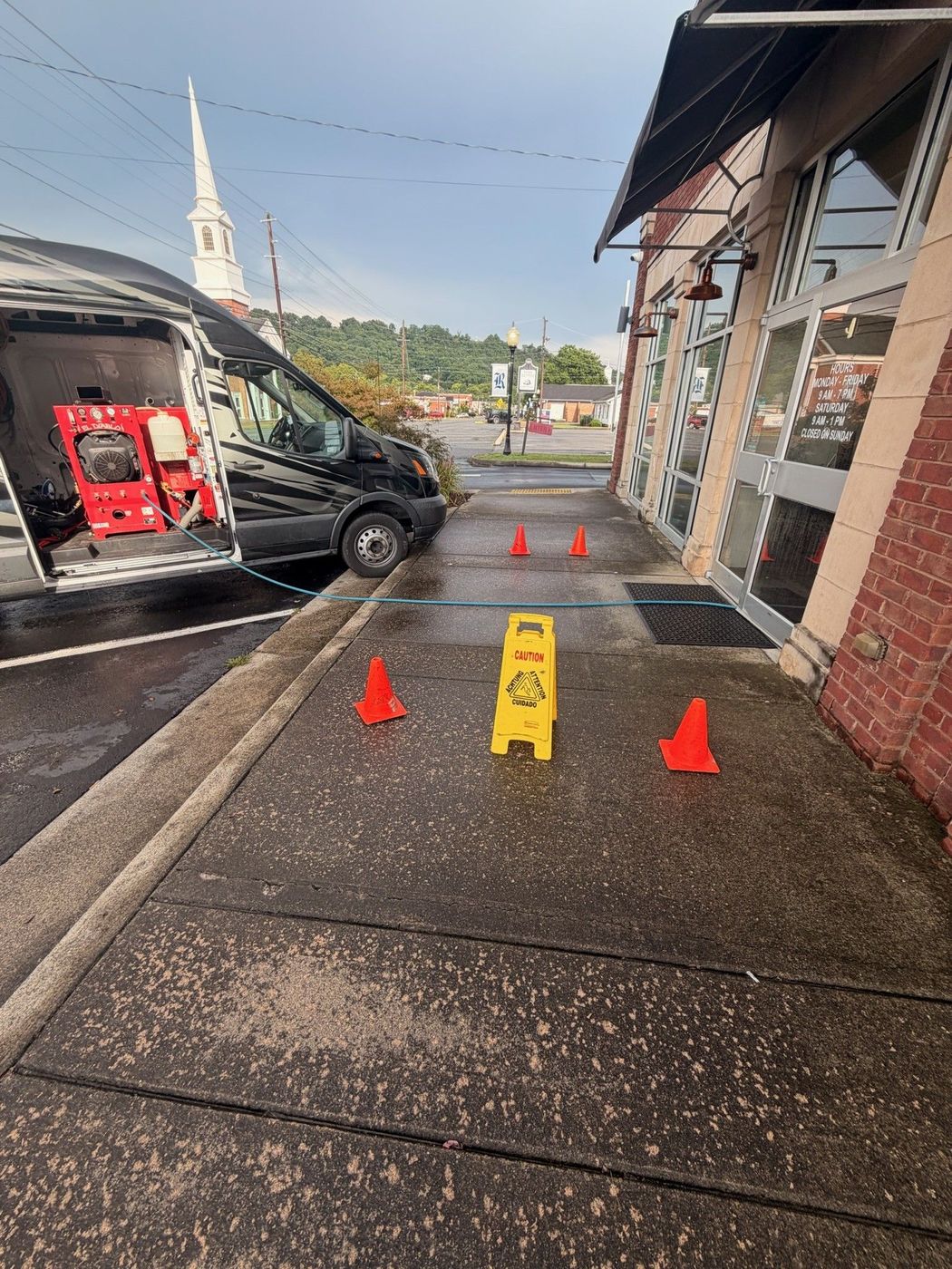 Orange cones and a yellow caution sign blocking a wet sidewalk beside a black van and building.