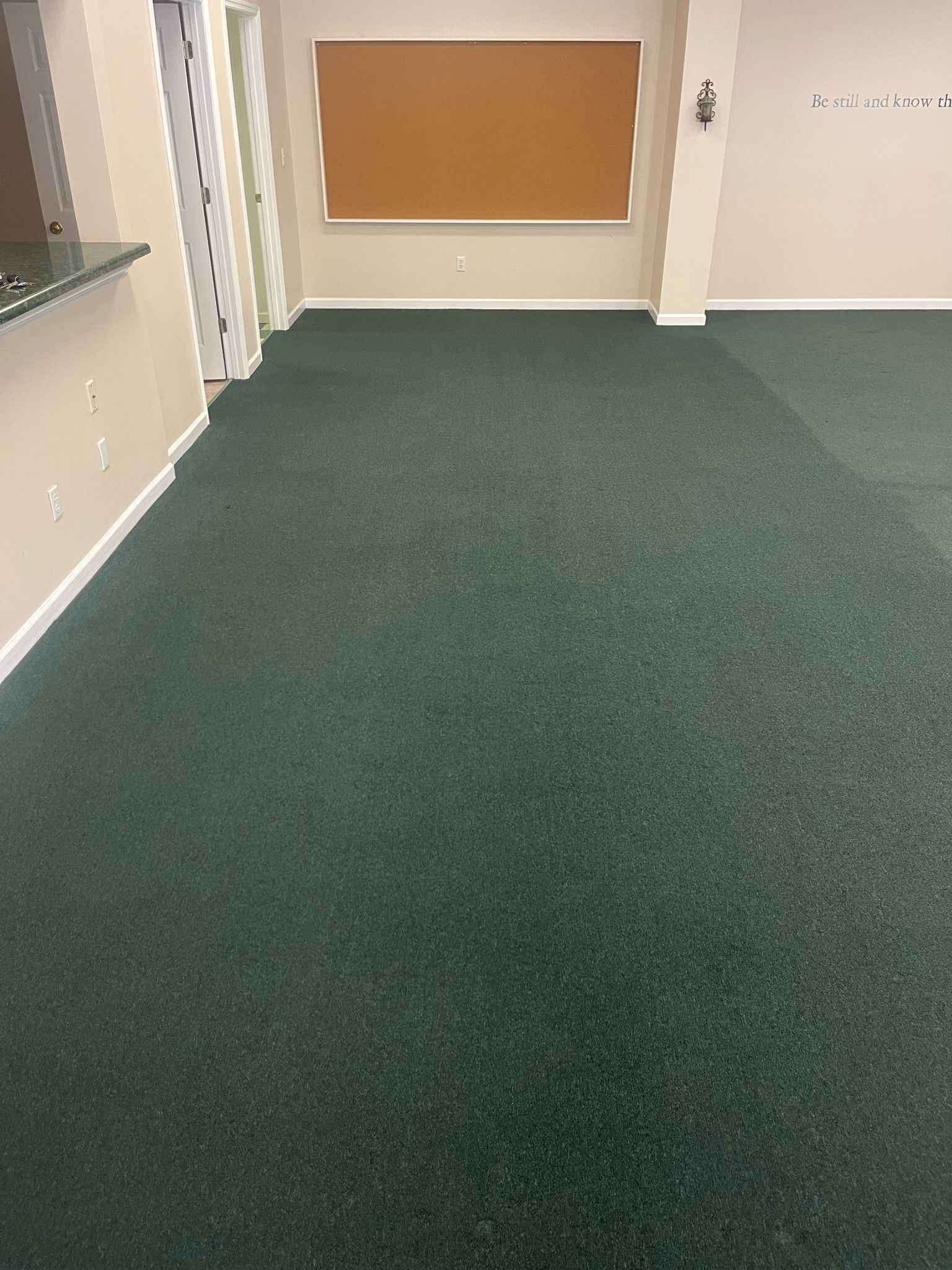 Empty hallway with green carpet, beige walls, and a bulletin board on the far wall.