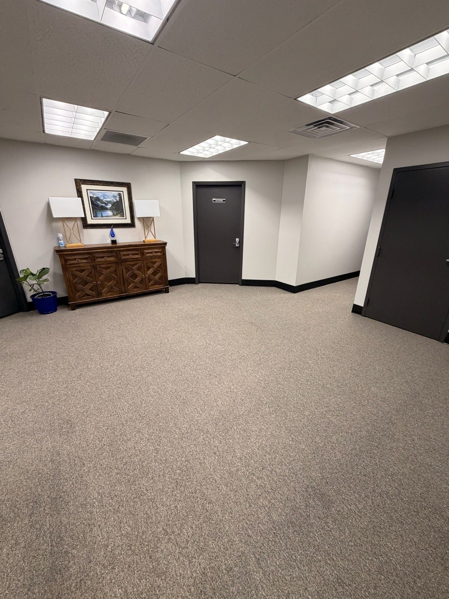 Empty office lobby with beige carpet, white walls, dark doors, and a wooden cabinet beneath a framed picture