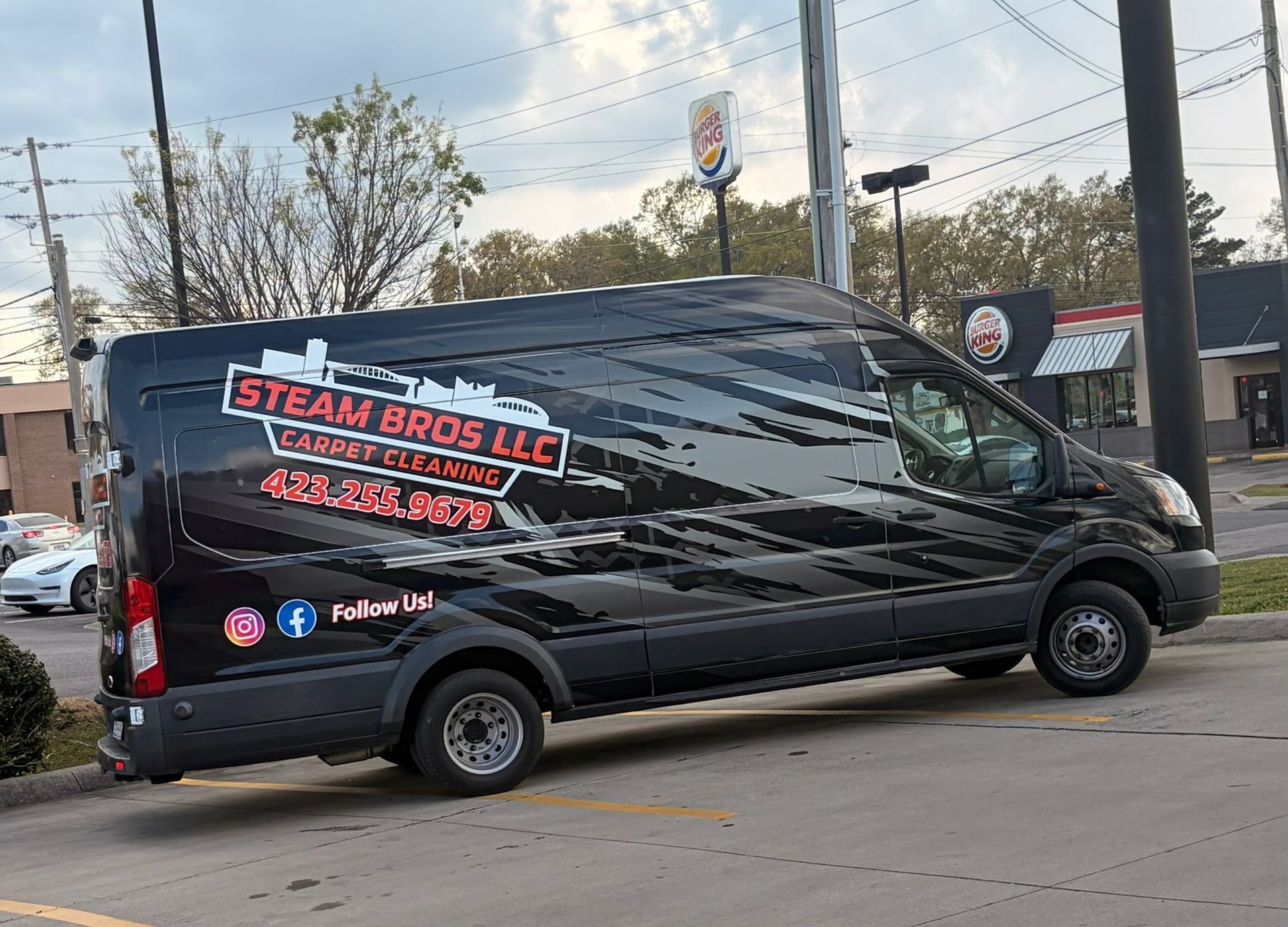 Black Steam Bros LLC van with logo parked on a street near utility poles and storefronts