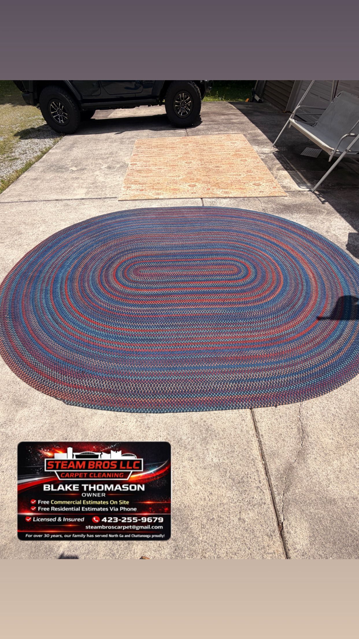 Red and blue oval rug on a concrete driveway beside a parked vehicle, with a small sign at lower left