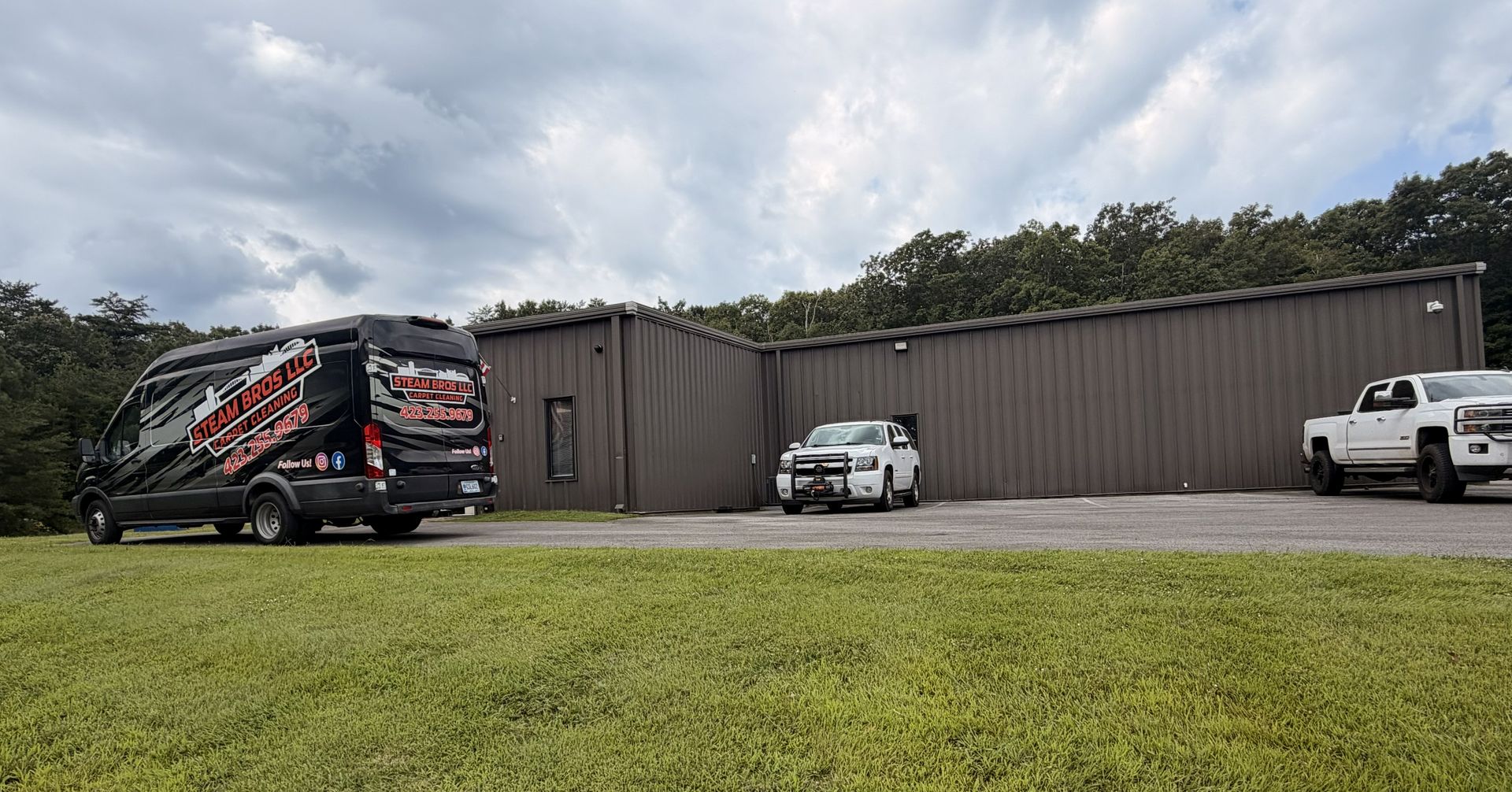 Parked vans and trucks beside a long brown metal building under cloudy skies.