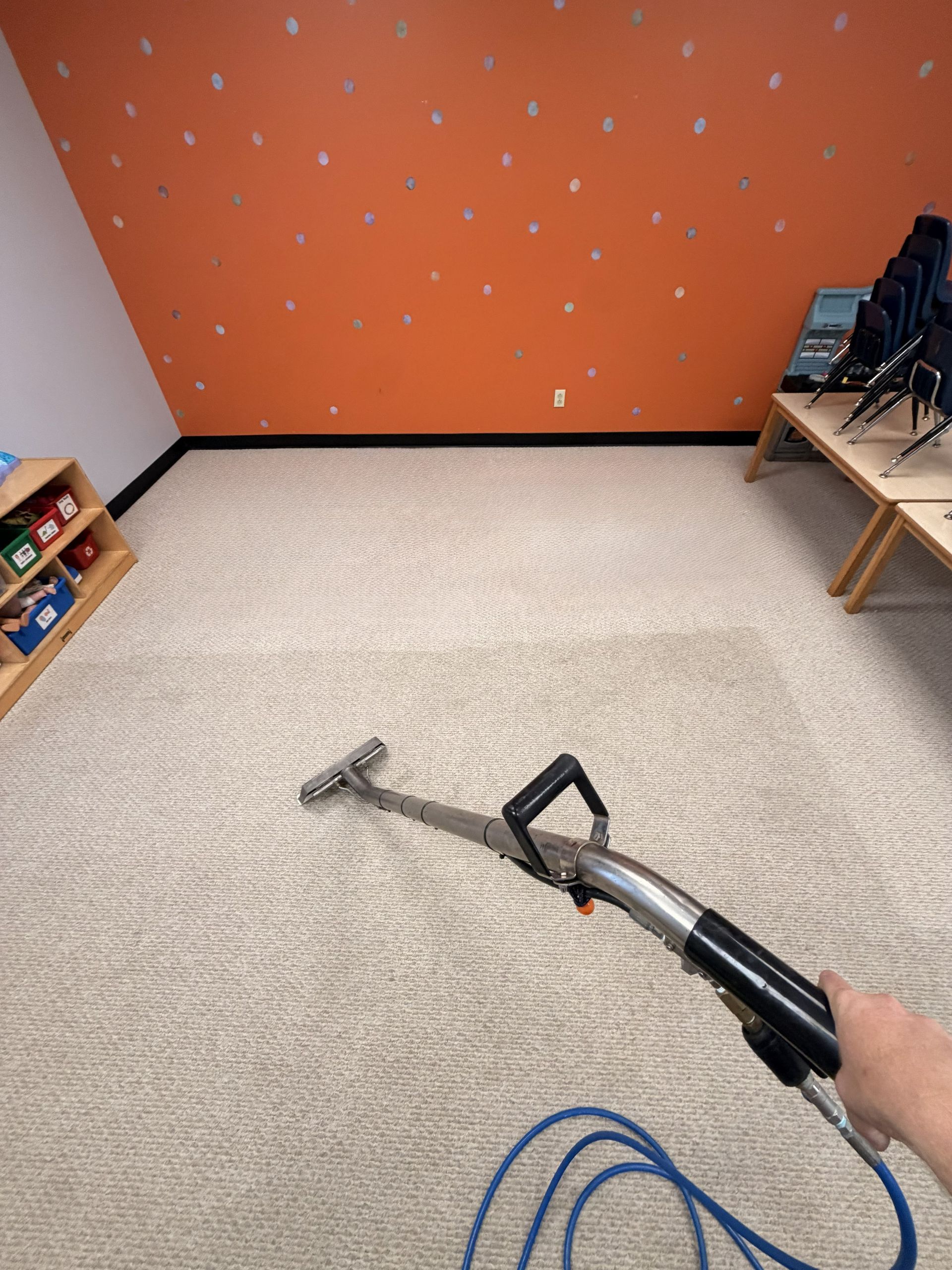 Carpet cleaning in a small room with orange wall; wand vacuuming beige carpet under a wood bench.