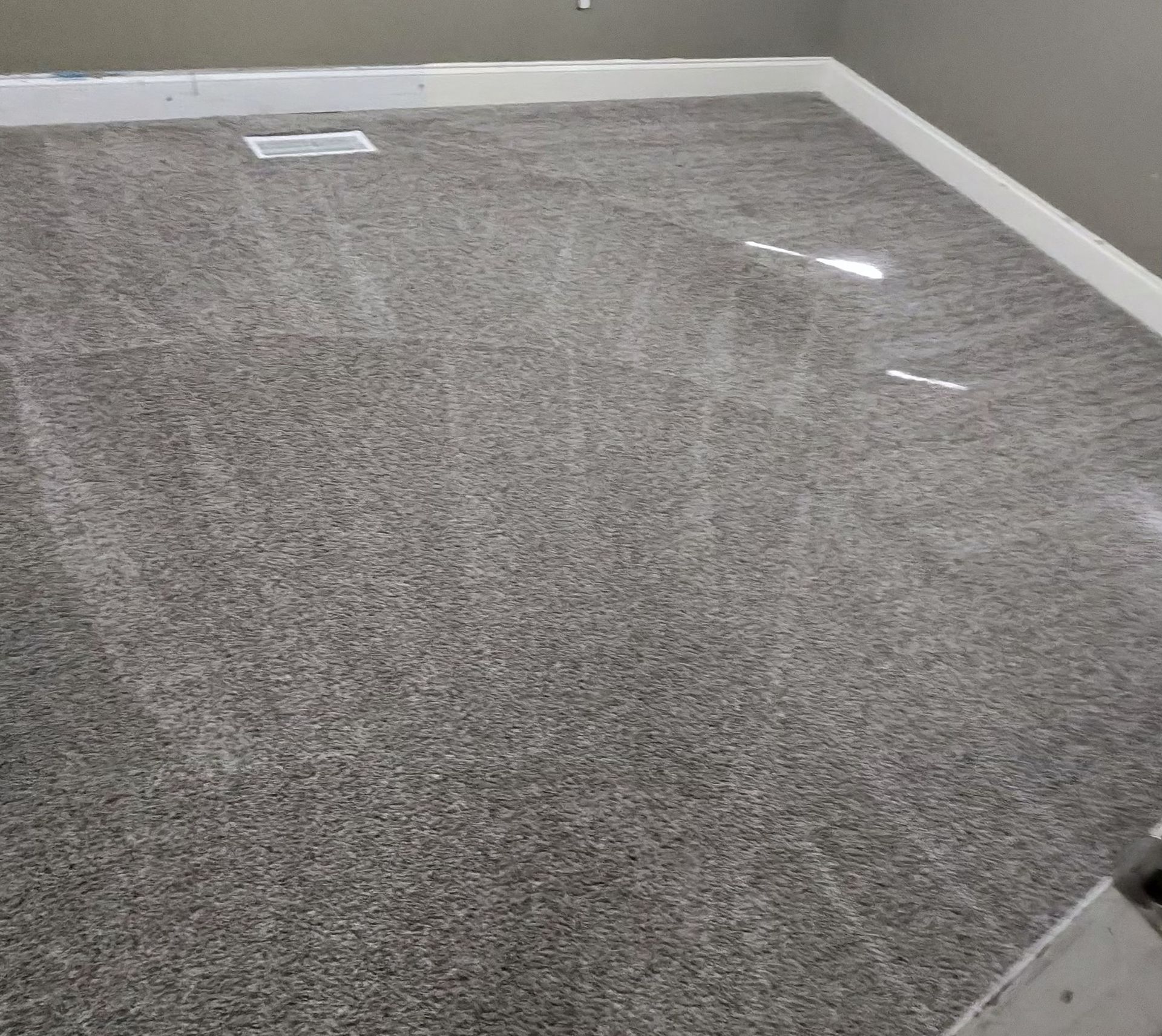 Empty gray carpeted room with white baseboards and two floor vents near the back wall