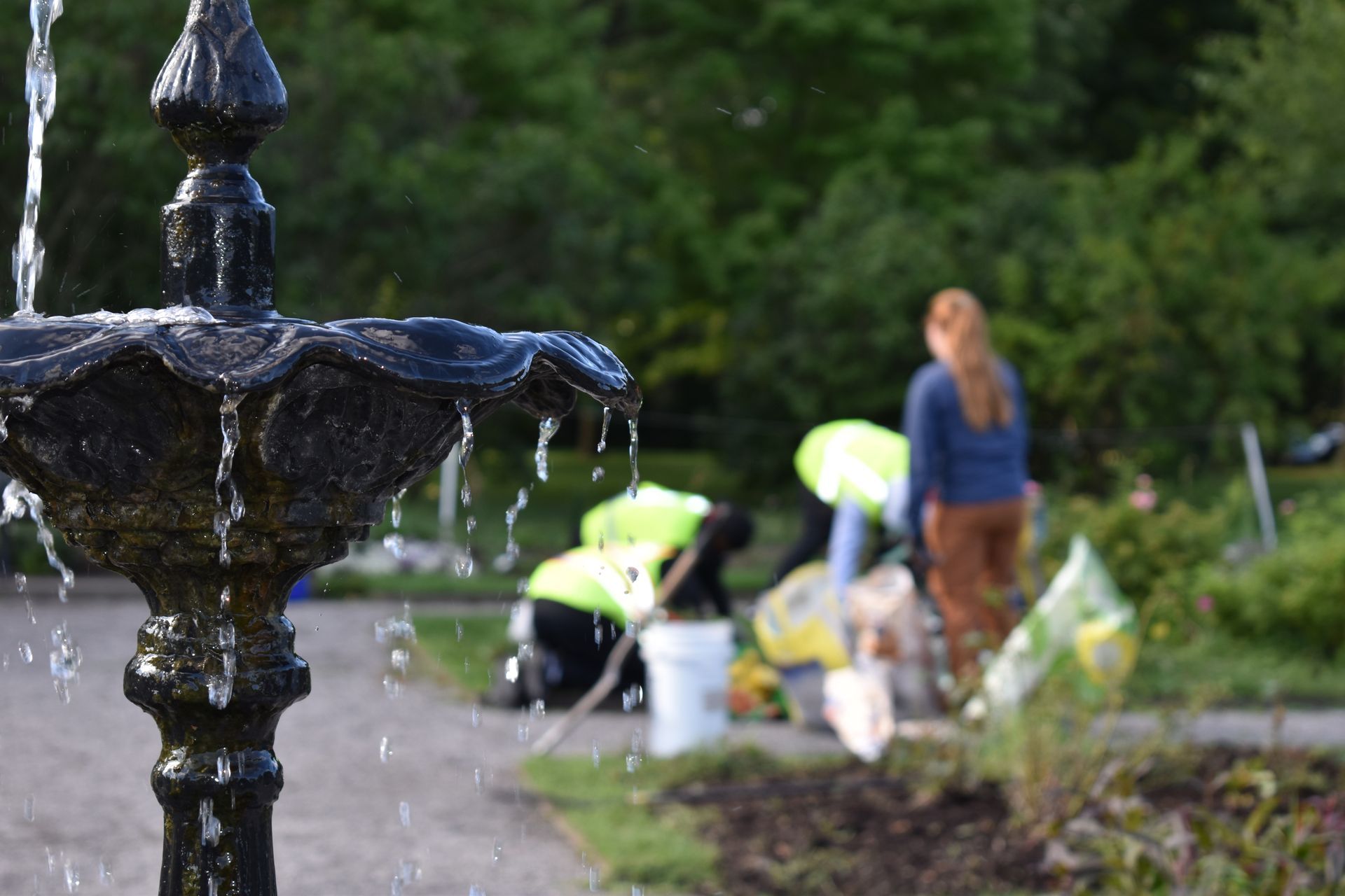 Black fountain in foreground with people gardening in blurred background near a path