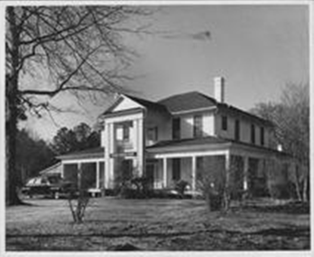 Black-and-white photo of a two-story house with a front porch, trees, and a car in the yard