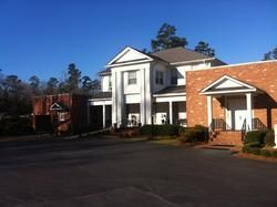 White colonial-style building with a red brick wing beside an empty parking lot on a sunny day