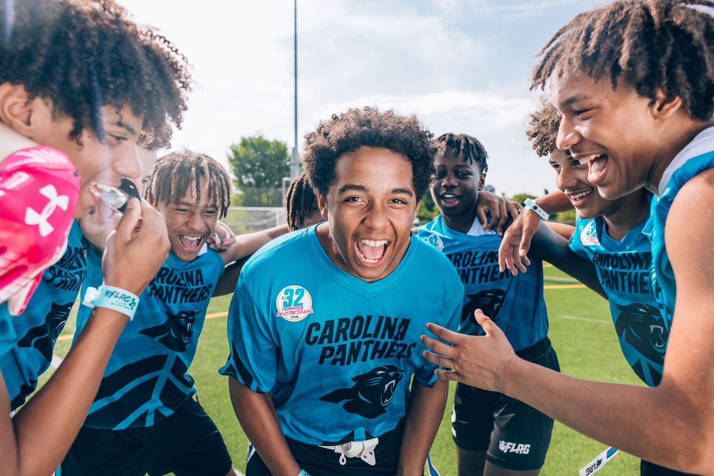 Football players huddle around a football, reaching hands together, blue sky backdrop.