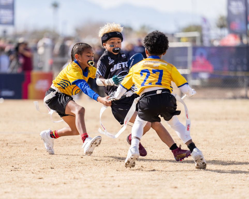 Football players in action on a grassy field; one runs with the ball, pursued by two opponents.