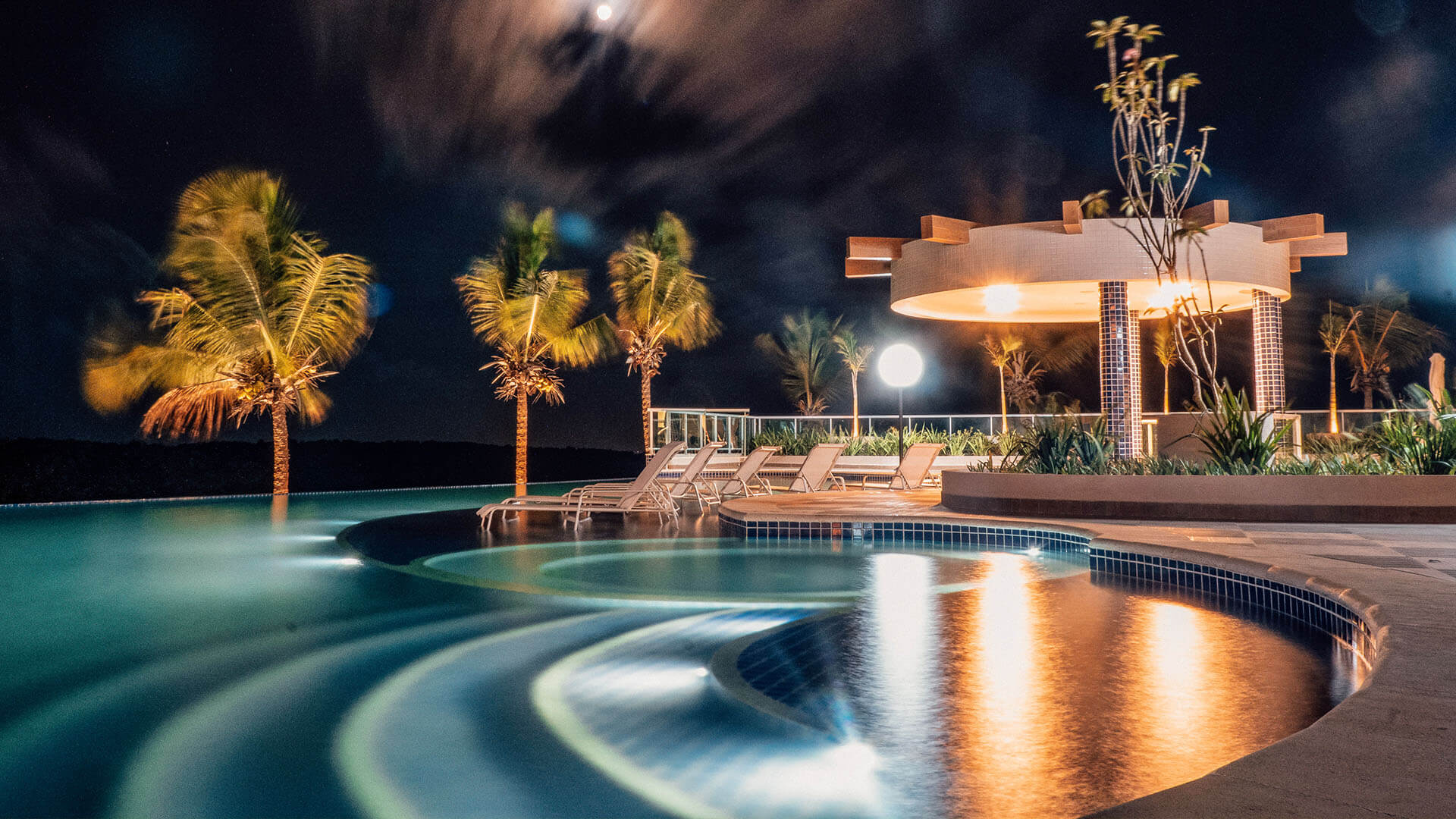 Uma grande piscina é iluminada à noite com palmeiras ao fundo.