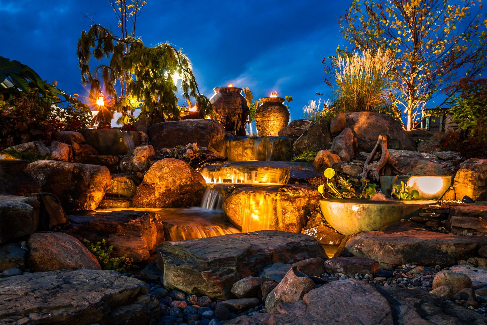Illuminated garden waterfall with rocks and warm lights under a deep blue evening sky