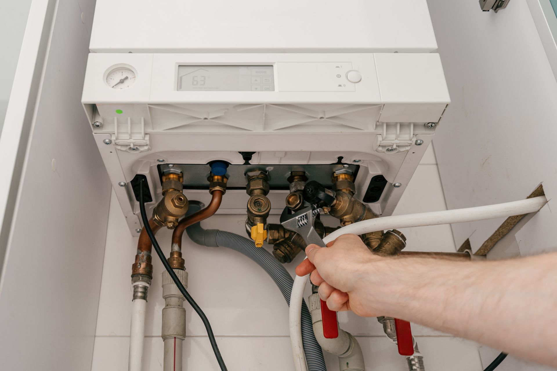 Person holding a red hose beneath an open wall-mounted boiler with exposed pipes and valves
