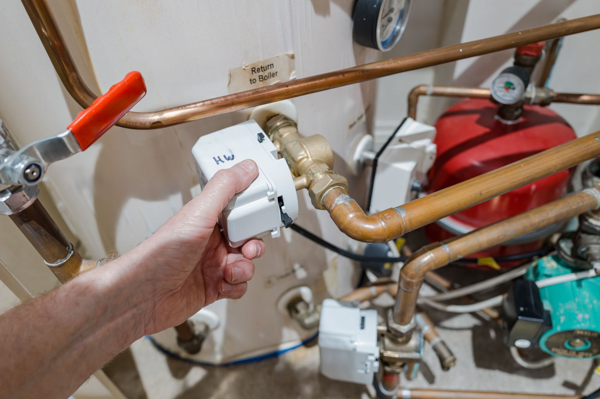 Hand holding a white valve controller on copper pipes beside a boiler and red expansion tank