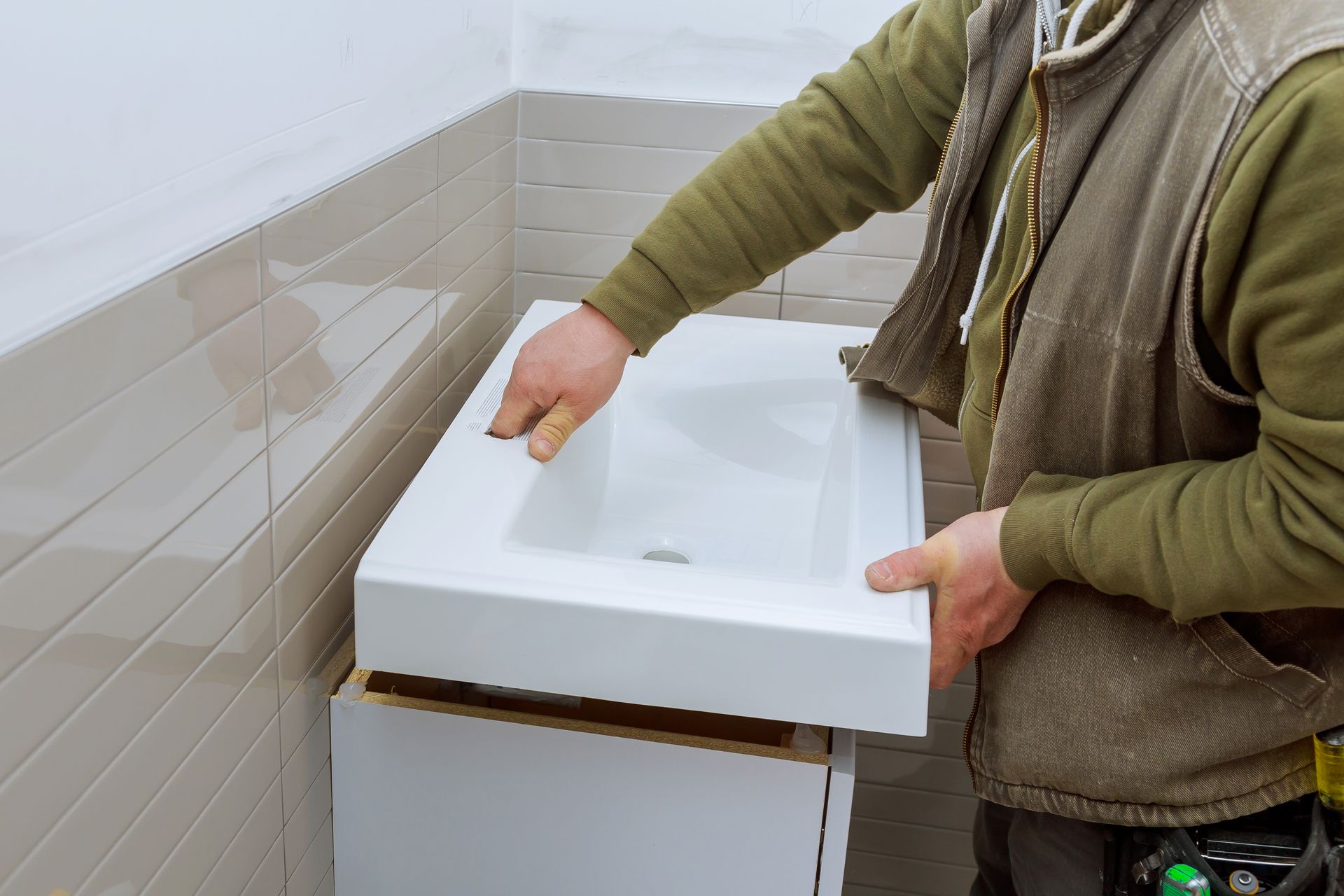 Person installing a white bathroom sink on a tiled wall with tools in hand