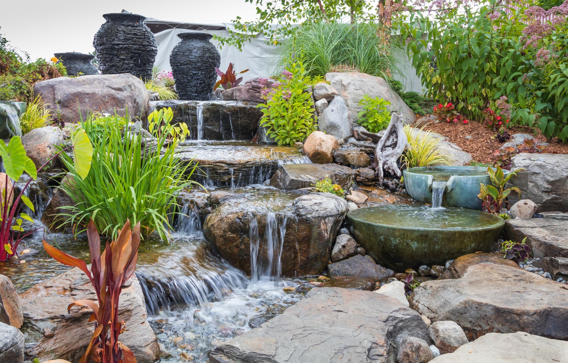 Tiered backyard waterfall with rocks, plants, and a small pond flowing over stone steps Tiered backyard waterfall with rocks, plants, and a small pond flowing over stone steps