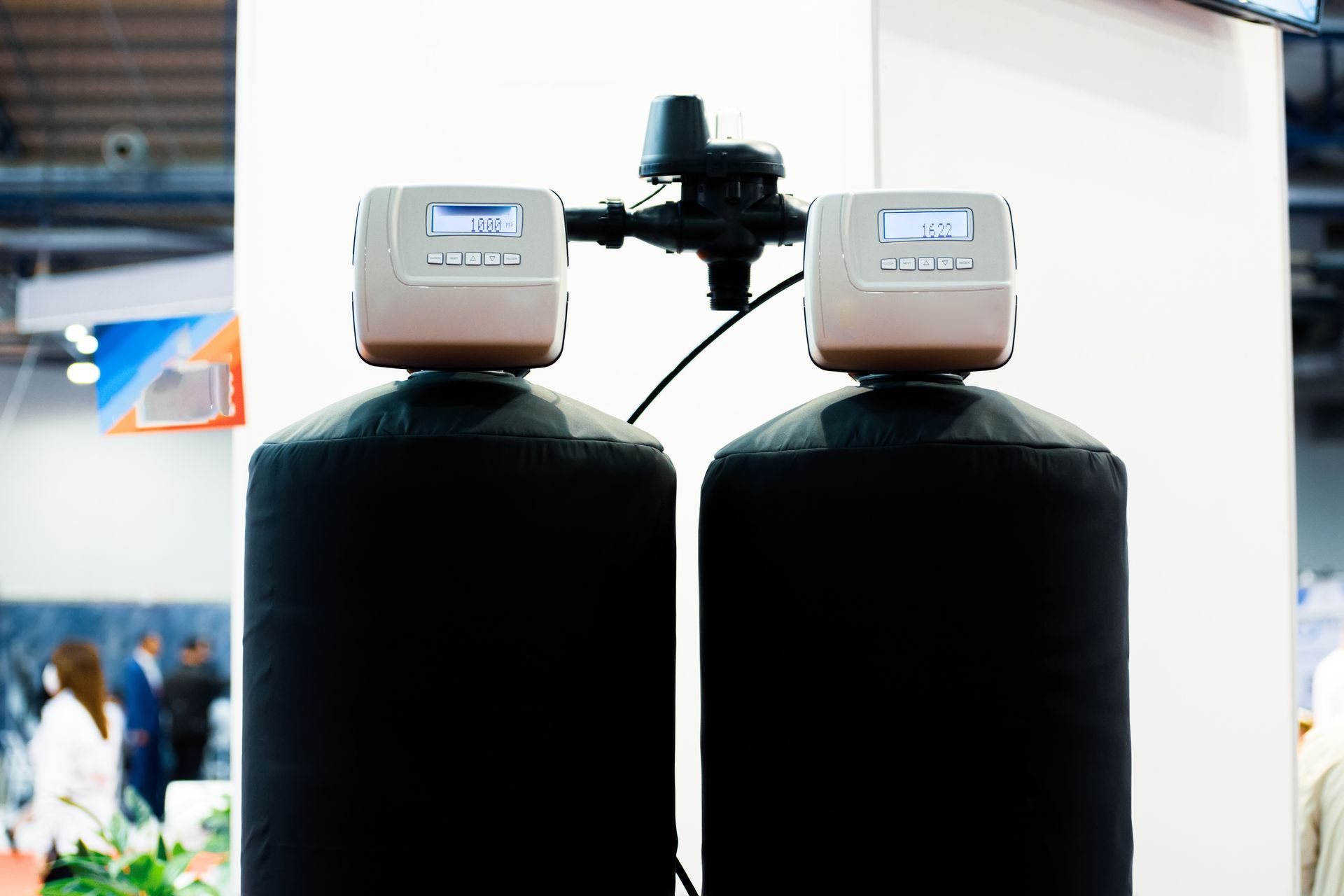 Two black cylindrical tanks with beige control units on top, set indoors at a trade show display. Two black cylindrical tanks with beige control units on top, set indoors at a trade show display.