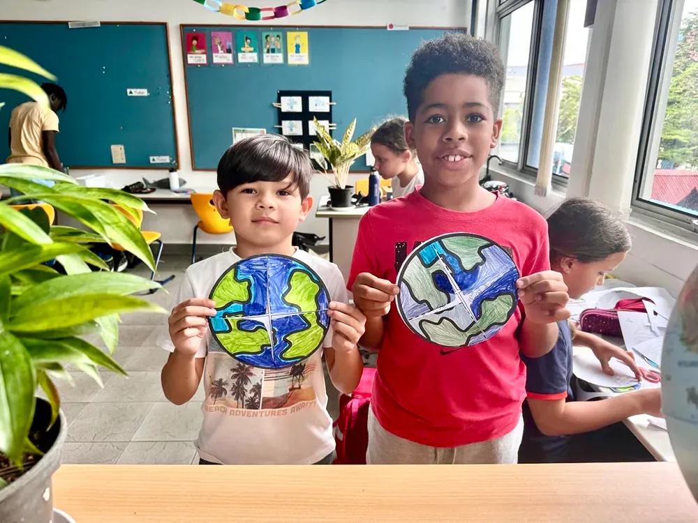 Two young boys are holding up their drawings of the earth in a classroom.
