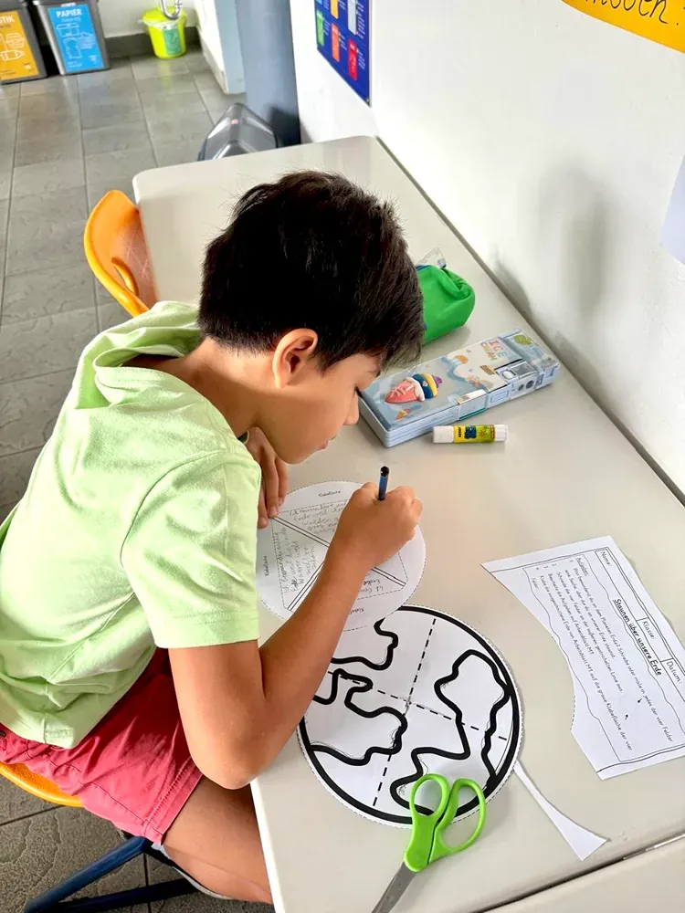 A young boy is sitting at a table drawing a picture of the earth.