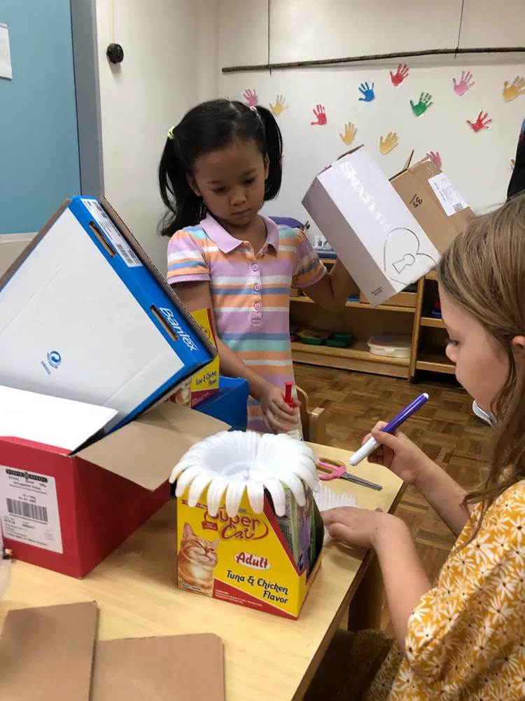 Two young girls are playing with cardboard boxes in a classroom.