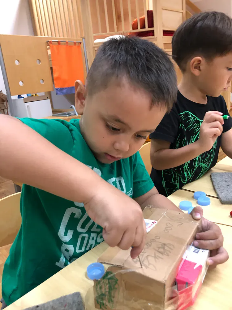 Two young boys are sitting at a table playing with a cardboard box.
