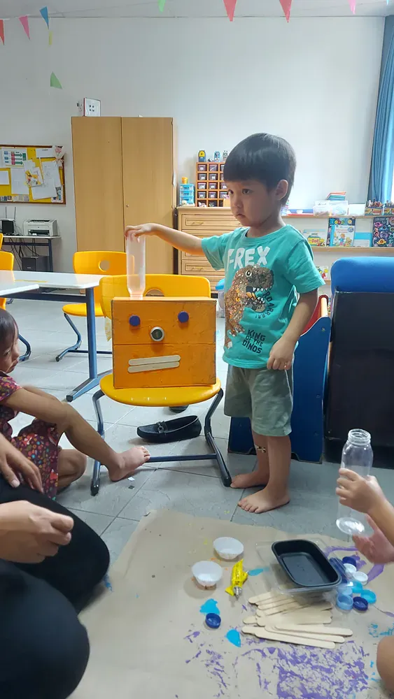 A young boy is standing in front of a box with a face on it in a classroom.