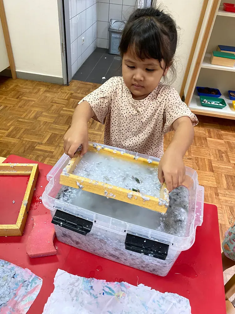 A little girl is sitting at a table playing with a box of shredded paper.