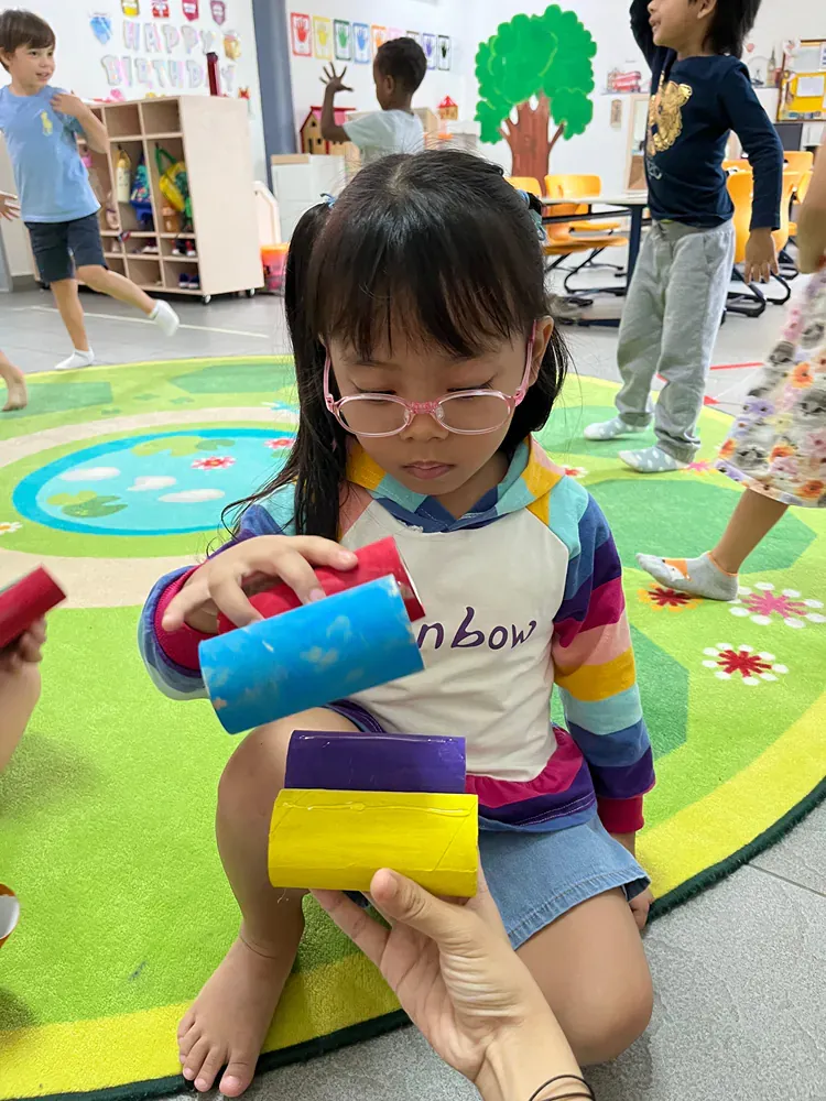 A little girl wearing glasses is playing with toilet paper rolls in a classroom.