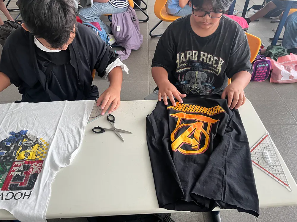A boy wearing a hard rock shirt is cutting a shirt with scissors