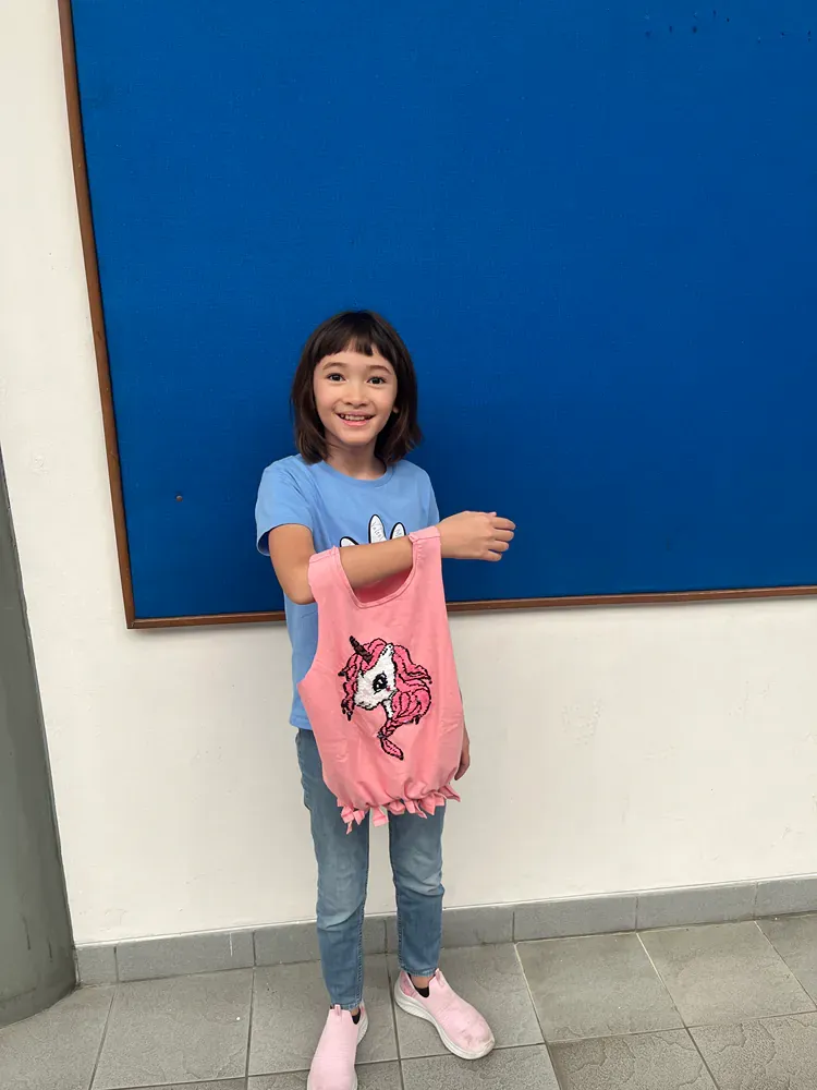 A little girl is standing in front of a blue board holding a pink shirt with a unicorn on it.