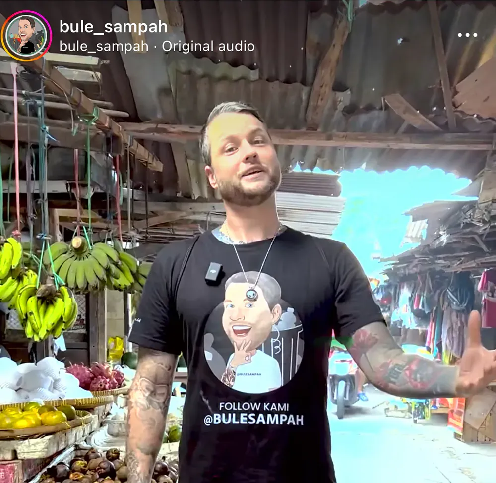 A man wearing a black t-shirt with a picture of a man on it is standing in front of a fruit stand.